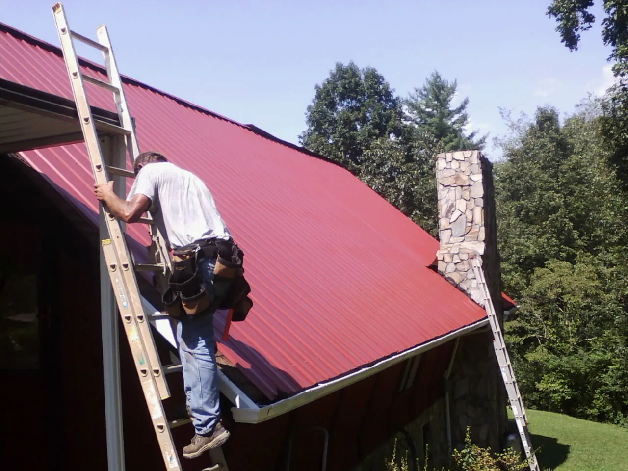 Roofer on a ladder beside a red metal roof and stone chimney — Soffit & Fascia Repair by Roofing Brothers in Glenn